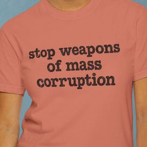 May include: A coral-coloured short-sleeve t-shirt featuring the black text "stop weapons of mass corruption" printed in a bold, slightly distressed font across the chest. The garment has a classic crew neckline and a standard, relaxed fit.
