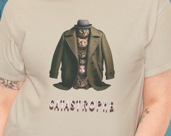 May include: Beige t-shirt featuring a whimsical design of cats stacked inside a green trench coat and a small hat. The word "CATASTROPHE" is printed in a maroon color below the graphic. The shirt is made of a soft material.