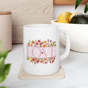 Peut inclure: Une tasse en céramique blanche avec un motif floral rose et blanc et le mot "LOVE" en lettres roses. La tasse a une anse et est posée sur un dessous-de-tasse beige.