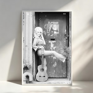 Puede incluir: Impresión enmarcada en blanco y negro que presenta a una mujer en denim apoyada contra una puerta, sosteniendo una guitarra. La puerta tiene un cartel que dice "THE GREENHOUSE DANCE ENSEMBLE". La imagen es un retrato, con la mujer sonriendo a la cámara.
