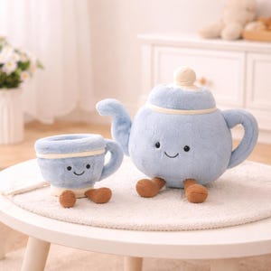 Puede incluir: Un juego de tetera y taza de peluche azul claro con caras sonrientes y pies marrones. La tetera tiene tapa y boquilla, mientras que la taza tiene asa. El conjunto está diseñado para parecer un personaje lindo y amigable.