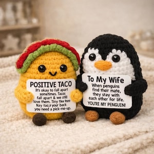 May include: Two crocheted amigurumi figures: a yellow taco with a red and green shell and a black and white penguin. Each holds a sign. The taco's sign reads "POSITIVE TACO". The penguin's sign reads "To My Wife".