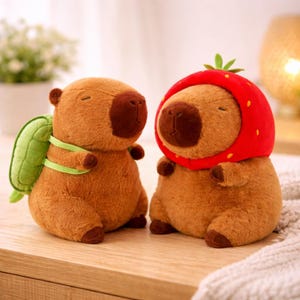 May include: Two plush capybaras. One wears a green turtle shell backpack, the other a red strawberry hat. Both are brown with dark brown noses and feet. The plush toys are soft and cuddly, perfect for children or collectors.