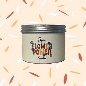 May include: A round container with a silver lid and a cream label. The label reads "Pepper's FLOWER POWER Sprinkles" in colourful, retro lettering, with flower and star graphics. The background is a light peach colour with scattered leaf shapes.