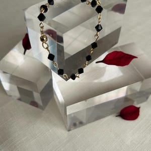 Puede incluir: Una delicada pulsera con cuentas negras facetadas ensartadas en una cadena dorada. La pulsera se exhibe sobre un bloque geom&eacute;trico transparente, con hojas rojas esparcidas. El cierre es un cierre magn&eacute;tico dorado.