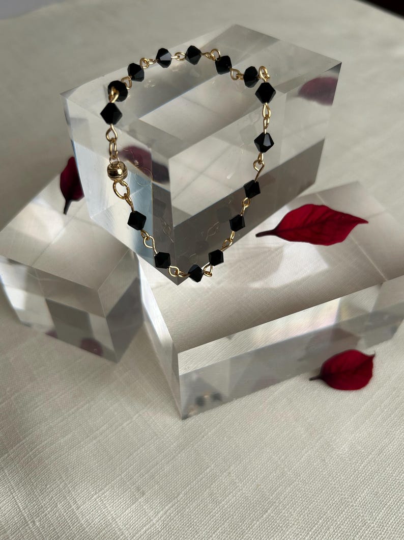 Puede incluir: Una delicada pulsera con cuentas negras facetadas ensartadas en una cadena dorada. La pulsera se exhibe sobre un bloque geom&eacute;trico transparente, con hojas rojas esparcidas. El cierre es un cierre magn&eacute;tico dorado.