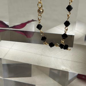 Puede incluir: Un delicado collar con cuentas negras facetadas ensartadas en una cadena dorada. El collar tiene un cierre magn&eacute;tico y se exhibe sobre una superficie clara y geom&eacute;trica. El fondo incluye un toque de rojo.