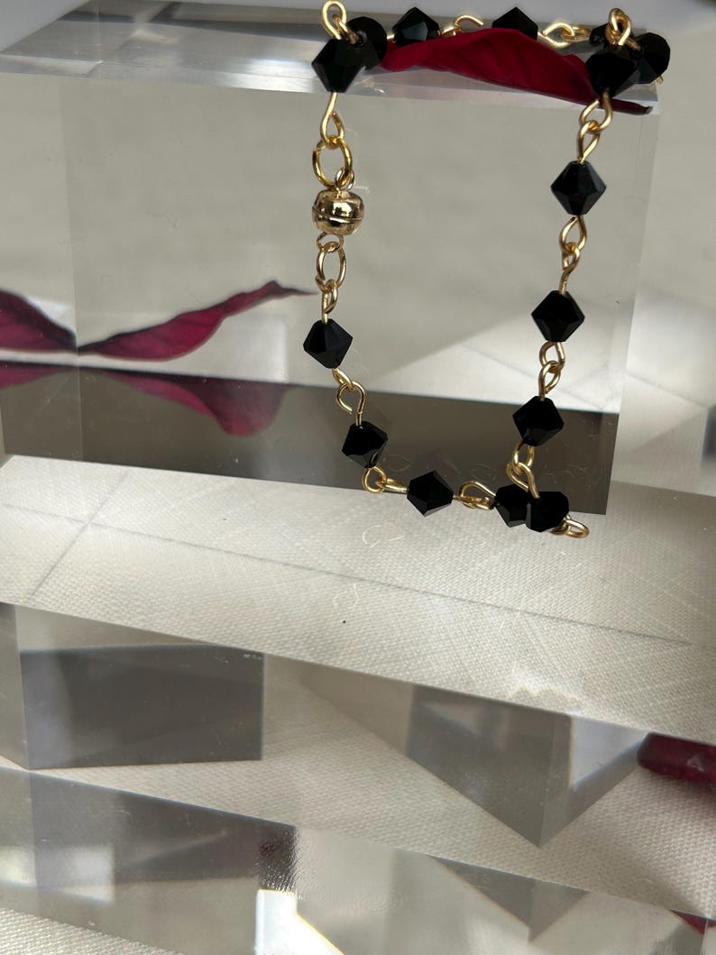 Puede incluir: Un delicado collar con cuentas negras facetadas ensartadas en una cadena dorada. El collar tiene un cierre magn&eacute;tico y se exhibe sobre una superficie clara y geom&eacute;trica. El fondo incluye un toque de rojo.