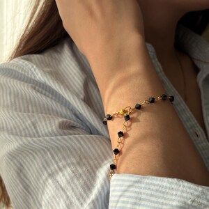 Puede incluir: Un brazalete dorado con cuentas cuadradas negras se lleva en la mu&ntilde;eca. El brazalete tiene un dise&ntilde;o delicado y sencillo. La persona lleva una camisa a rayas azul claro y blanca.