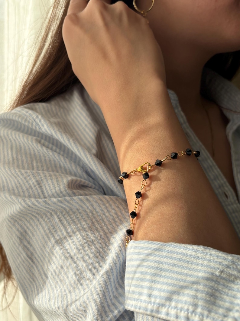 Puede incluir: Un brazalete dorado con cuentas cuadradas negras se lleva en la mu&ntilde;eca. El brazalete tiene un dise&ntilde;o delicado y sencillo. La persona lleva una camisa a rayas azul claro y blanca.