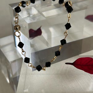 Puede incluir: Una delicada pulsera dorada con cuentas negras de corte cuadrado. Las cuentas est&aacute;n ensartadas con alambre dorado, y la pulsera tiene un cierre magn&eacute;tico. La joya se exhibe sobre una superficie transparente y geom&eacute;trica.