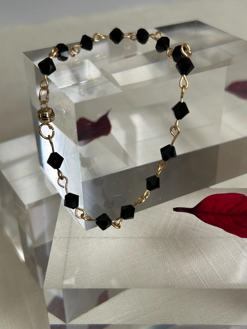 Puede incluir: Una delicada pulsera dorada con cuentas negras de corte cuadrado. Las cuentas est&aacute;n ensartadas con alambre dorado, y la pulsera tiene un cierre magn&eacute;tico. La joya se exhibe sobre una superficie transparente y geom&eacute;trica.