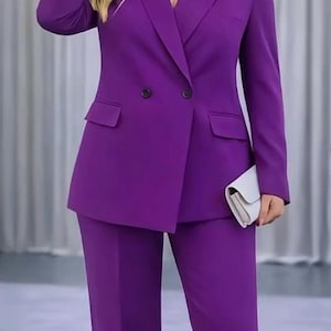 May include: A vibrant purple pantsuit featuring a double-breasted blazer with two front pockets and matching trousers. The outfit is paired with a white clutch. The image includes the text "Mushis Pakistan".