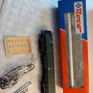 May include: A model train car in olive green with black detailing, alongside its packaging. The box is blue and red with the "Roco" logo. Additional accessories and paperwork are also visible, including a small tube of glue.