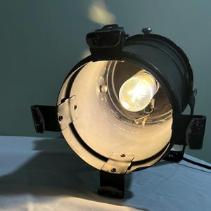 Puede incluir: Un foco negro de estilo vintage con una bombilla brillante e iluminada. El foco tiene un cuerpo cilíndrico con soportes ajustables y un cable de alimentación. La luz está colocada sobre una superficie blanca, proyectando una sombra.