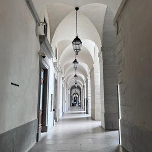 Peut inclure: Un long couloir voûté avec une série de lanternes noires suspendues. Les murs et les colonnes sont blancs, et le sol est en pierre. La perspective crée une sensation de profondeur, attirant le regard le long du couloir.