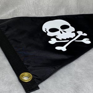 May include: A black triangular flag featuring a white skull and crossbones graphic. The left side has a thick black reinforced strip with two brass-colored metal grommets for mounting. The fabric has a smooth, slightly reflective finish.