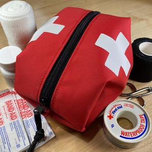 May include: A red first aid kit bag with a white cross symbol and a black zipper, surrounded by medical supplies including a white bandage roll, a white pill bottle, three Band-Aid packets, a black wrap, metal scissors, and waterproof tape.
