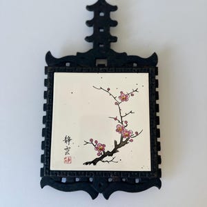 May include: A square ceramic tile featuring a hand-painted plum blossom branch with pink flowers on a white background, set within a dark, ornate, carved wooden frame with a pagoda-style top handle and black Chinese calligraphy characters.