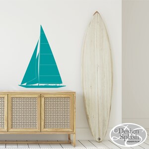 May include: Teal sailboat wall decal on a white wall with a wooden surfboard leaning against the wall. The decal is above a wooden cabinet with a woven rattan front.