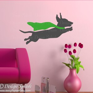 May include: A gray silhouette of a dachshund wearing a green cape, flying through the air. The dachshund is facing right and has its tongue sticking out. The background is a light pink wall.