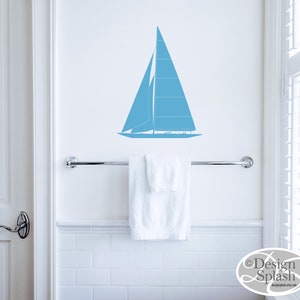 May include: A blue sailboat wall decal in a bathroom setting. The decal is above a white towel hanging on a chrome towel bar. The bathroom has white subway tile walls.