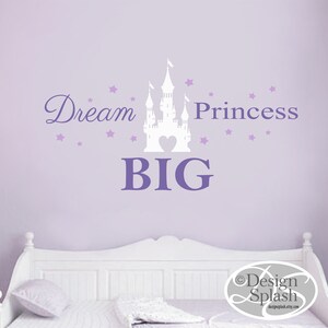 May include: Purple wall with a white castle silhouette and the text "Dream Big Princess" in purple lettering. There are small purple stars scattered around the castle and text. The wall is behind a white bed with a white pillow.