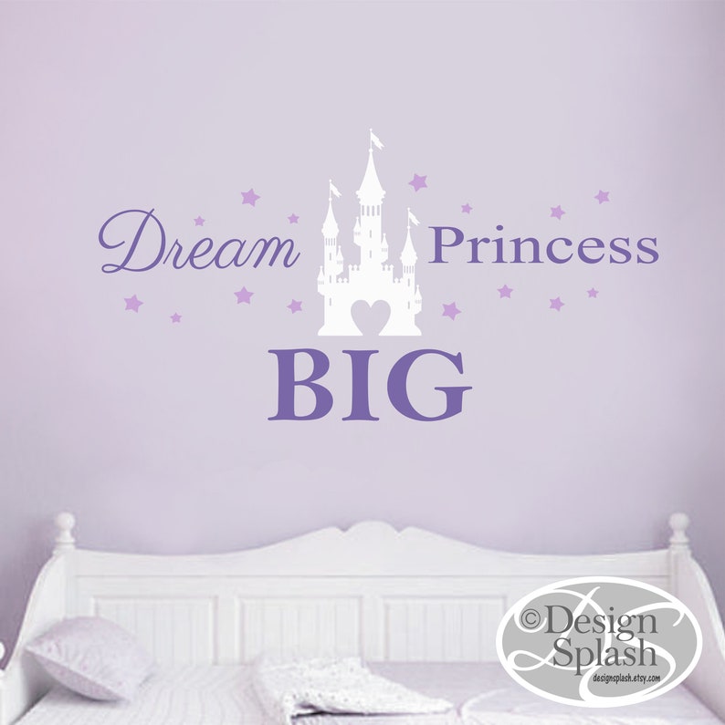 May include: Purple wall with a white castle silhouette and the text "Dream Big Princess" in purple lettering. There are small purple stars scattered around the castle and text. The wall is behind a white bed with a white pillow.