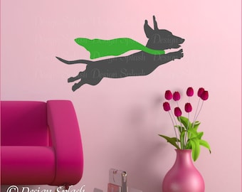 May include: A gray silhouette of a dachshund wearing a green cape, flying through the air. The dachshund is facing right and has its tongue sticking out. The background is a light pink wall.
