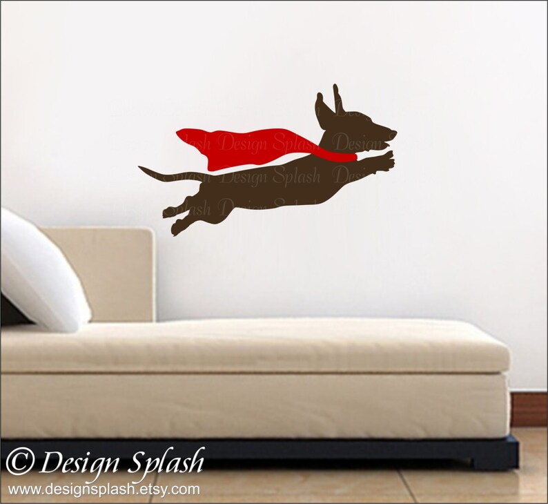 May include: A brown dachshund wearing a red cape, flying through the air. The dog is silhouetted against a white wall. The image is a graphic design for a wall decal.