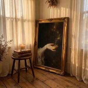 May include: A large, ornate gold framed painting leans against a wall. The painting depicts a hand reaching out to catch golden sparkles against a dark background. A small wooden stool holds stacked books and a lit candle.