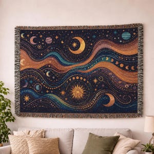 May include: A woven tapestry featuring a celestial design with moons, stars, and planets in shades of gold, blue, and orange. The tapestry has a dark blue background and a fringed edge. The design includes a large crescent moon and a sun.