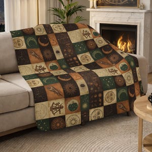 May include: A cozy throw blanket with a celestial design. The blanket features a patchwork pattern of squares in shades of brown, green, and beige. Each square displays illustrations of the sun, moon, stars, and other astronomical elements. The blanket is draped over a beige sofa.
