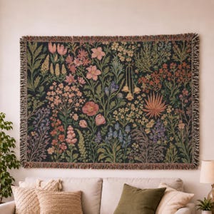 Peut inclure: Un plaid tissé rectangulaire présentant un fond sombre avec un motif botanique dense et coloré composé de diverses fleurs, feuilles et tiges dans des tons de rose, orange, bleu et vert, fini avec un bord décoratif à franges.