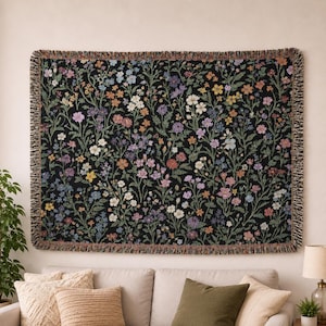 May include: A rectangular black tapestry with a floral pattern in shades of pink, purple, yellow, and white. The tapestry has a fringed border and is hanging on a wall above a sofa. The floral design features various flowers and green leaves.