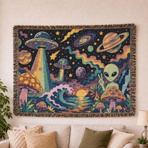 Peut inclure: Un plaid tissé représentant une scène spatiale vibrante avec un grand extraterrestre faisant un signe de paix, des champignons colorés, des soucoupes volantes, des planètes et des étoiles sur un fond sombre parsemé d'astres célestes.
