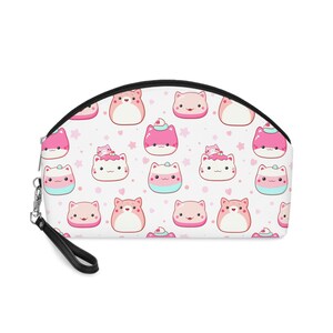 Op de afbeelding: Witte make-uptas met een zwarte ritssluiting en polsband. De tas heeft een herhalend patroon van cartoonkatten in roze, wit en blauw met steraccenten. De katten hebben verschillende ontwerpen, waaronder slagroom en glazuur.