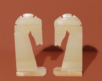 May include: A pair of white marble bookends shaped like horse heads. The bookends are facing each other and have a smooth, polished finish.