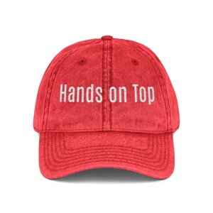 May include: A red, vintage-style baseball cap with the text "Hands on Top" embroidered in white. The cap has a curved brim and a button on top. The fabric has a slightly faded appearance.