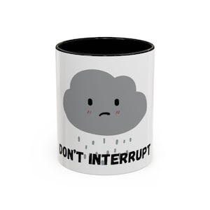 May include: White ceramic mug with a black interior and the words "DON'T INTERRUPT" in black. The mug features a gray cartoon cloud with a sad face and rain droplets.