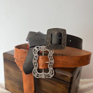 May include: Three leather belts with decorative buckles are displayed on a wooden box. The belts are in shades of brown and black. One belt has a silver filigree buckle, while another has a dark metal buckle with an ornate design. The third belt is a dark brown colour.