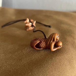 May include: A carved wooden netsuke pendant featuring a mermaid figure with long hair and a scaled tail, holding a small sphere. The brown wooden carving is attached to a thin black cord that loops through the figure, showcasing intricate details.