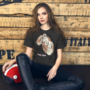 May include: Dark gray t-shirt featuring a tiger head graphic with the text "Wild Tiger" in pink. The tiger design is in shades of brown and white. The person is wearing black pants and is sitting next to a red and white helmet.