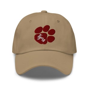May include: A tan baseball cap featuring a maroon paw print with the word "Tigers" embroidered in white script. The cap has a curved brim and a button on top, suitable for sports fans.