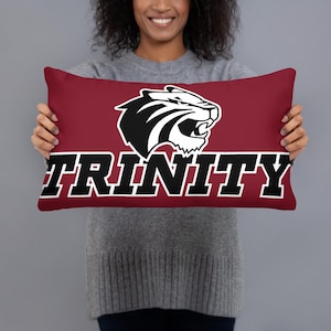 May include: A maroon rectangular pillow featuring a black and white tiger head logo above the word "TRINITY" in bold, black letters. The pillow is held by a person wearing a gray sweater.