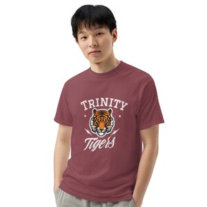 May include: A maroon t-shirt featuring the text "TRINITY Tigers" in white, with a tiger graphic in the center. The tiger is orange and black. The shirt is a short-sleeved, crew neck style.