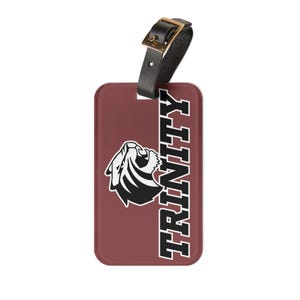 May include: A maroon luggage tag with the word "TRINITY" in black and white vertical text. The tag features a black and white graphic of a tiger head and a black leather strap with a gold buckle.