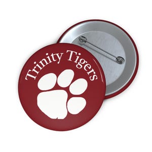 May include: A maroon button with the text "Trinity Tigers" in white, along with a white paw print. The button has a silver pin on the back and is a round shape.