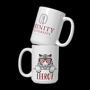 May include: Two white ceramic mugs stacked on top of each other. The top mug features the Trinity University logo in gray and maroon. The bottom mug has a tiger wearing red glasses and the name "LEEROY" in maroon.