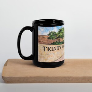 May include: Black ceramic mug featuring the text "TRINITY UNIVERSITY" in bold, white letters. The mug displays a scenic image of a campus building and trees under a blue sky. The mug is placed on a wooden surface.